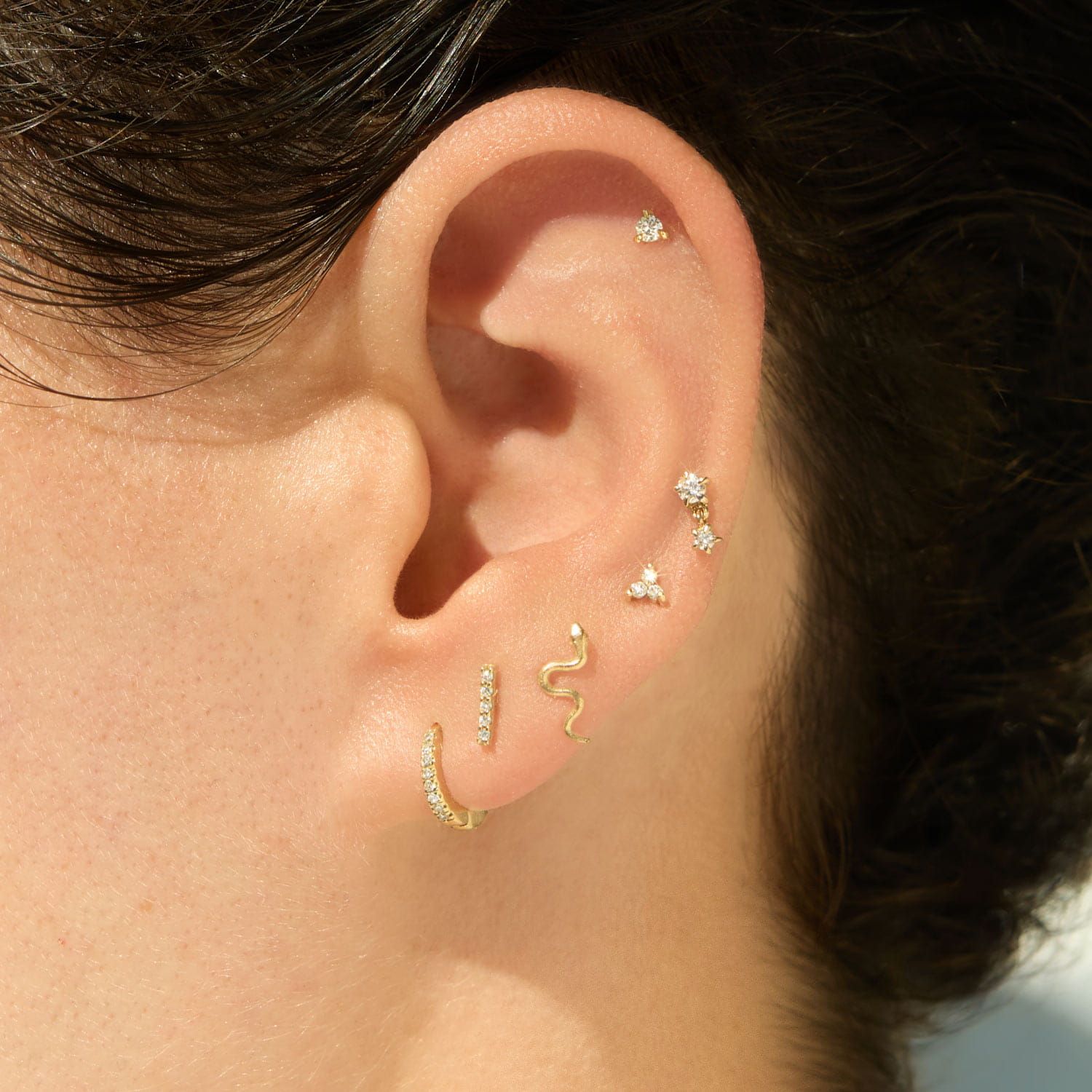 Close-up of an ear with multiple gold and gemstone piercings, including a snake-shaped stud and a stone-set hoop.
