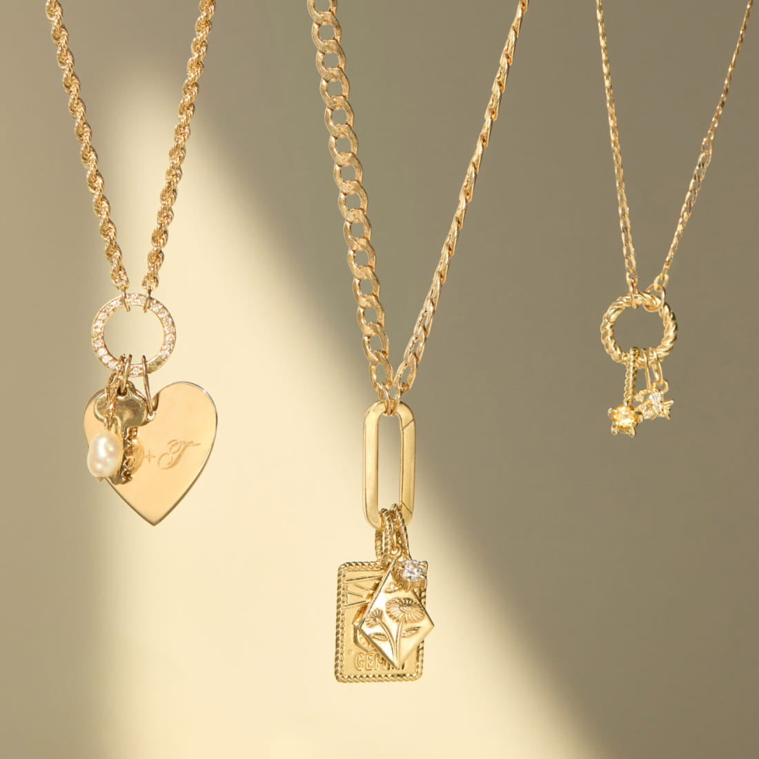 Three gold necklaces displayed on a neutral background, featuring a heart pendant, a layered oval charm, and a star-embellished circular charm.