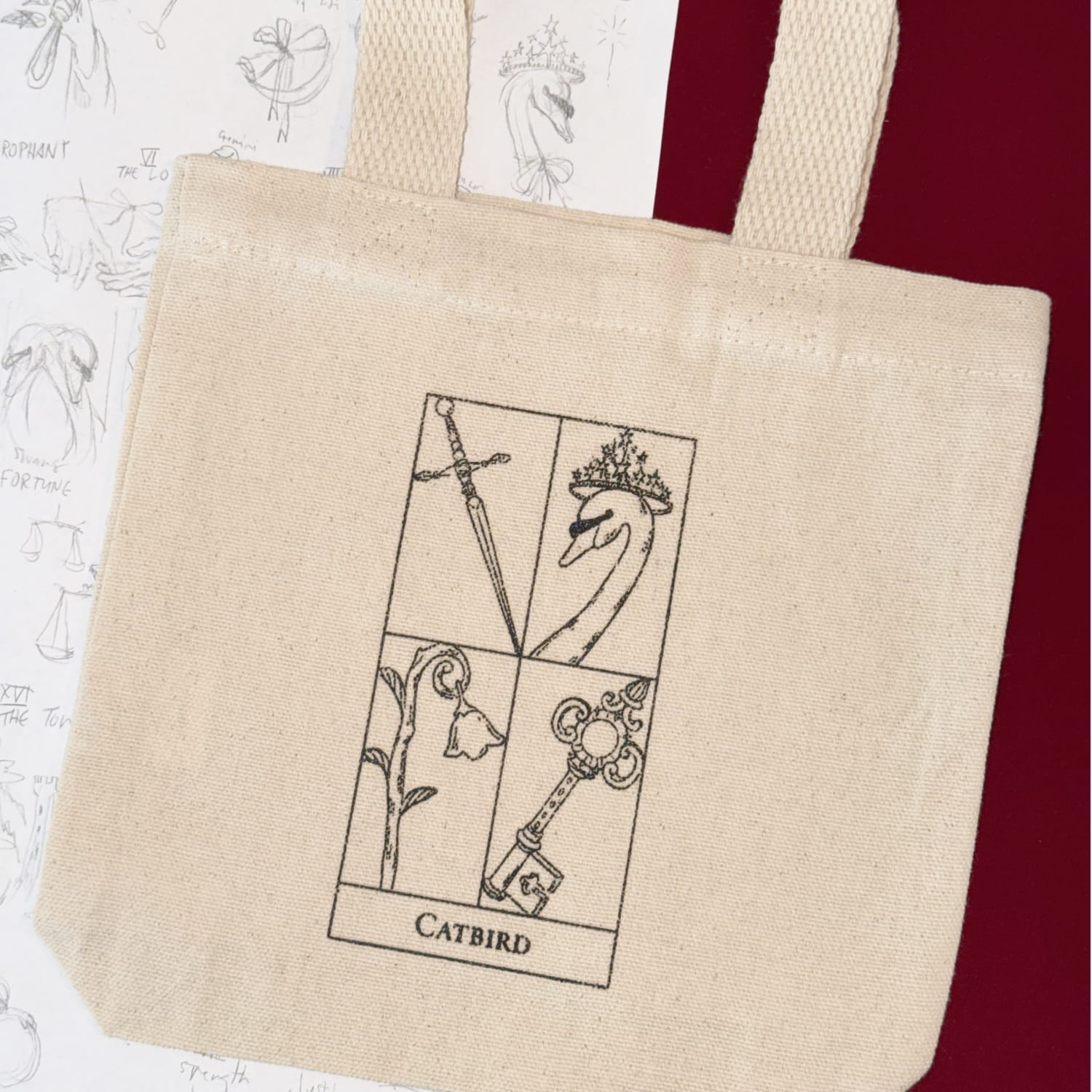 Beige Clairo canvas tote bag with a four-panel black illustration of a sword, crowned bird, flower, and ornate key, labeled &ldquo;CATBIRD.