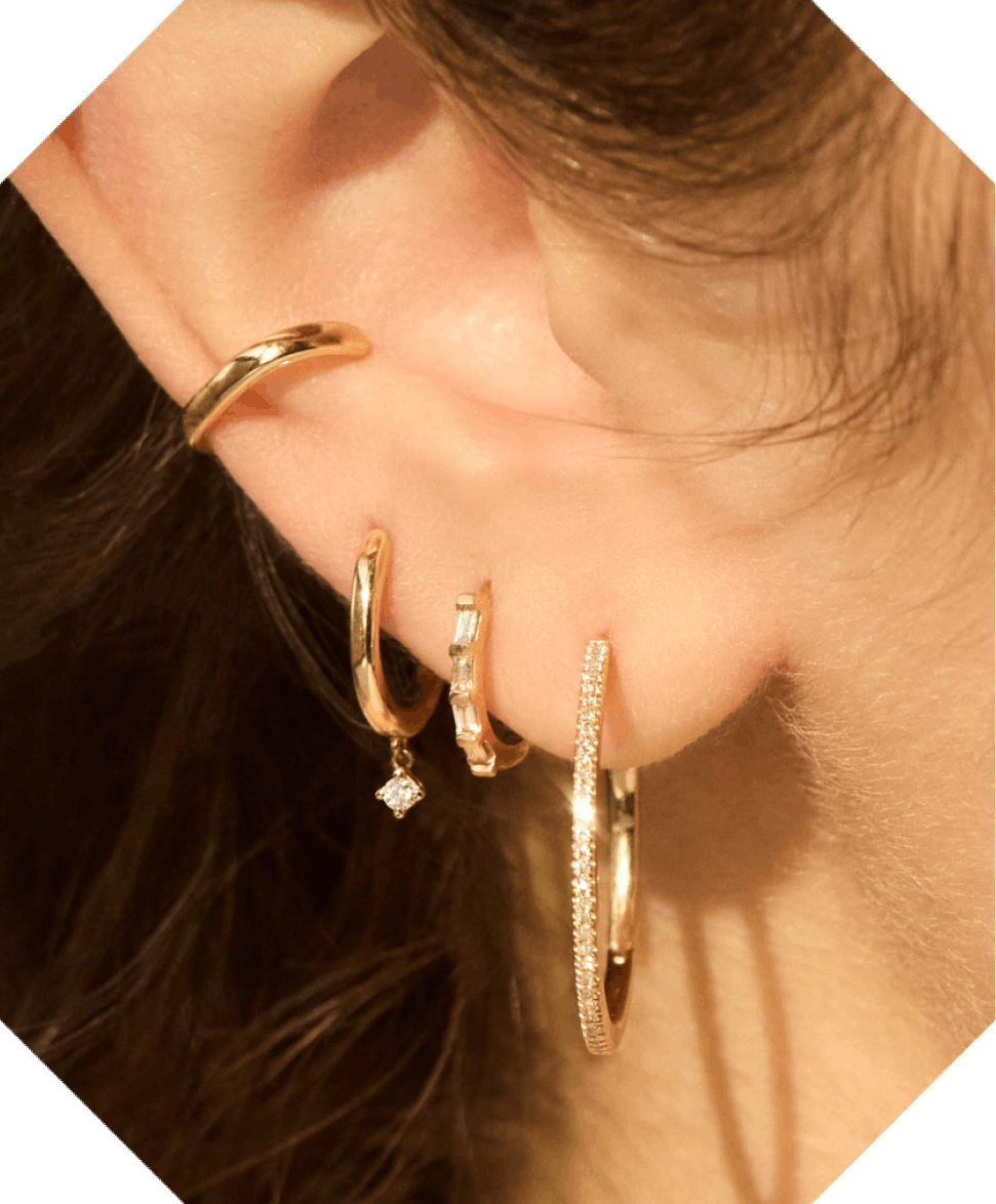 Close-up of a person's ear adorned with multiple gold hoop earrings, varying in size and texture, one with a small diamond charm, against a warm-toned background.