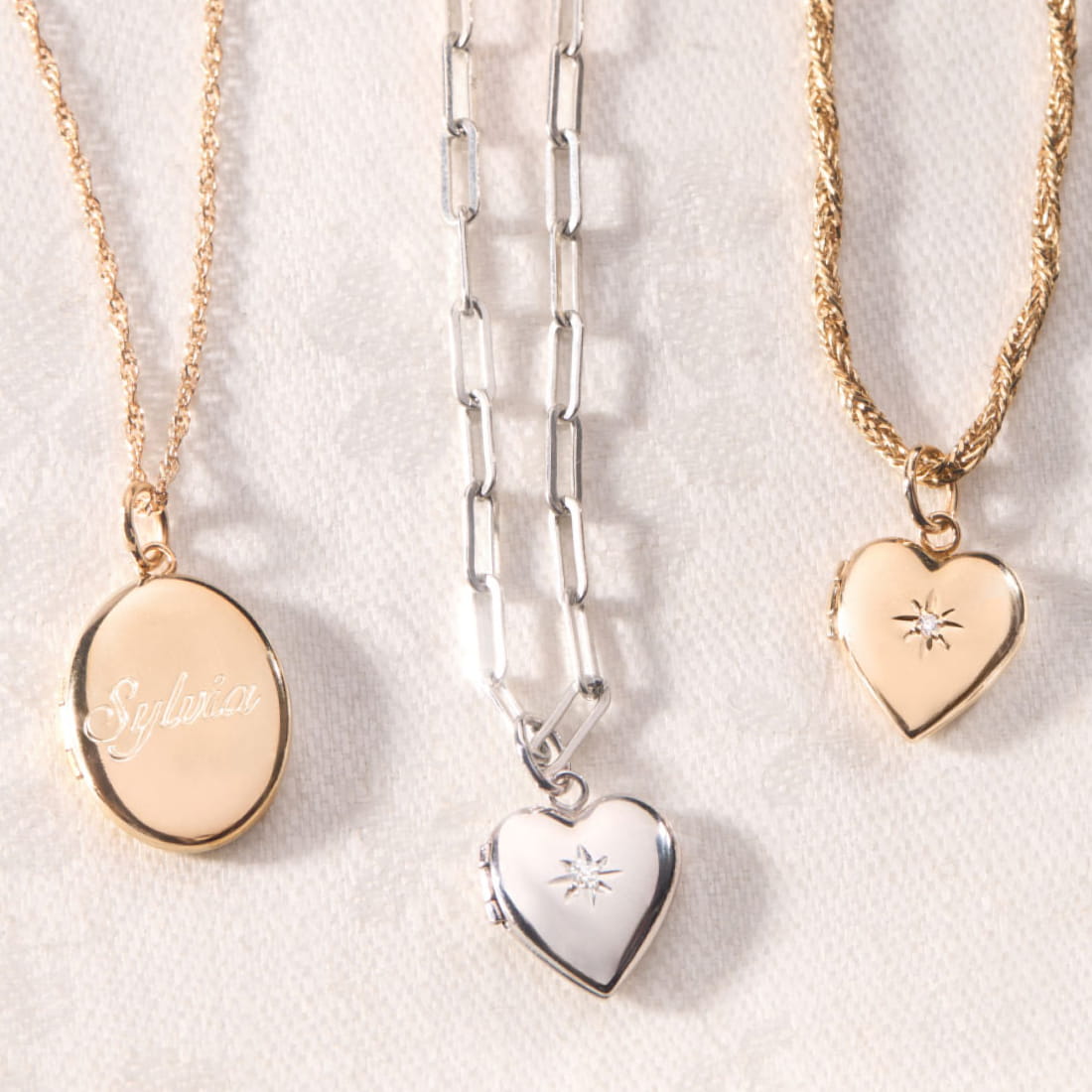 Three lockets on a white lace background: a gold oval locket with "Sylvia," a silver heart with star engraving, and a gold heart with star motif. 