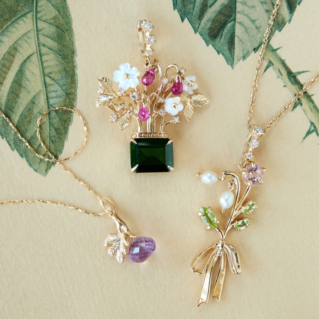 Three gold jewelry pendants on a light brown background: a flower bouquet pendant with green and pink gemstones, a purple droplet pendant with a leaf, and a slender bouquet featuring pearls and green accents.