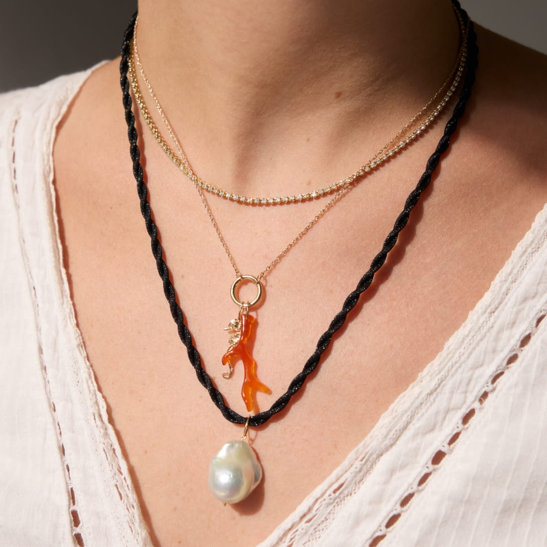 A close-up of a person wearing three layered necklaces. The top necklace is gold, the middle has a small gemstone, and the bottom features a large pearl.