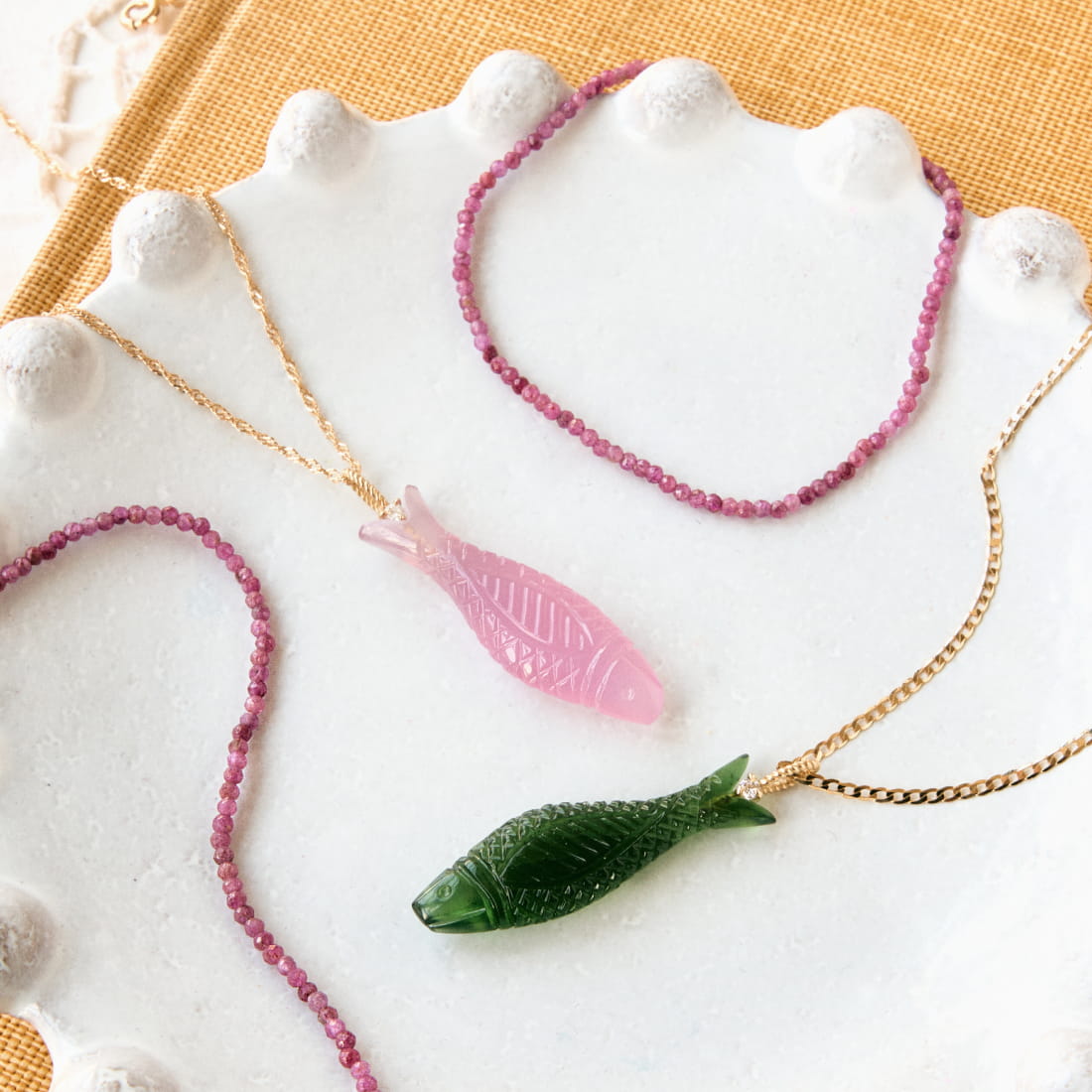 A white plate holds two fish-shaped pendants&mdash;one pink and one green&mdash;along with several pink bead necklaces, all set against a textured orange background