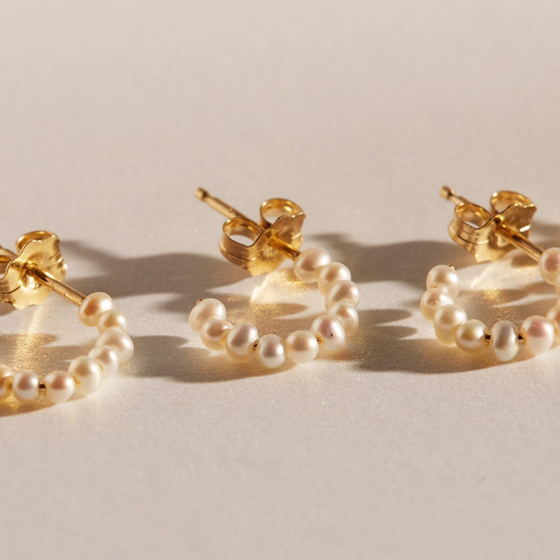 Three gold pearl hoop earrings arranged on a beige surface with warm lighting and shadows.