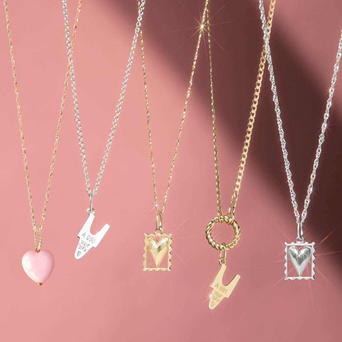 Five necklaces with heart-themed pendants and small tags hang against a pink background