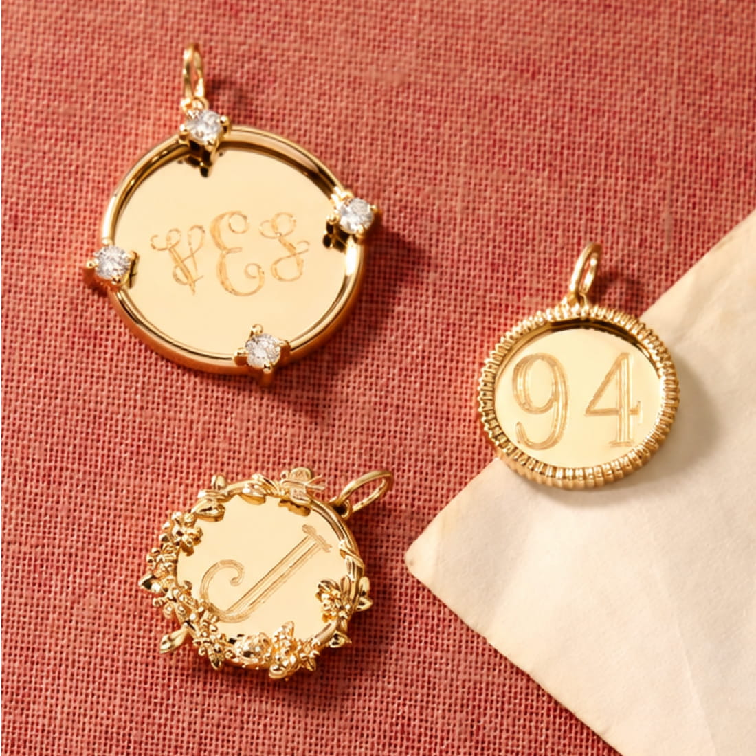 Three gold-toned engraved pendants arranged on rose-colored fabric, showing ornate initials and numbers.