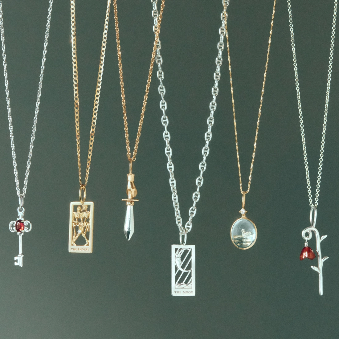 Six silver and gold necklaces with decorative pendants displayed against a dark green background, including &ldquo;THE LOVERS&rdquo; and &ldquo;THE MOON&rdquo; tarot-style pendants.