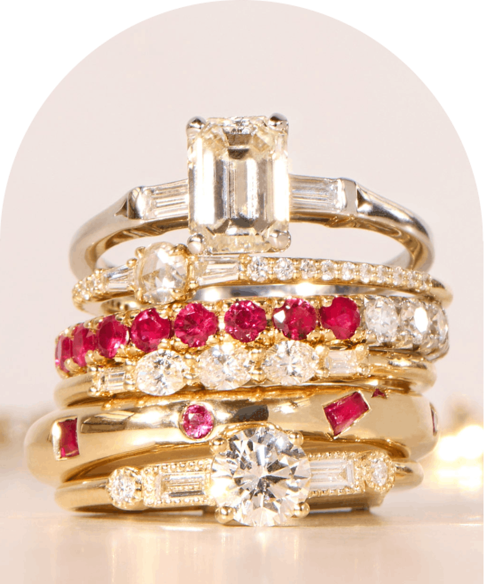 A stack of gold rings featuring various designs with diamonds and ruby gemstones, arranged in a vertical pile on a light-colored surface.