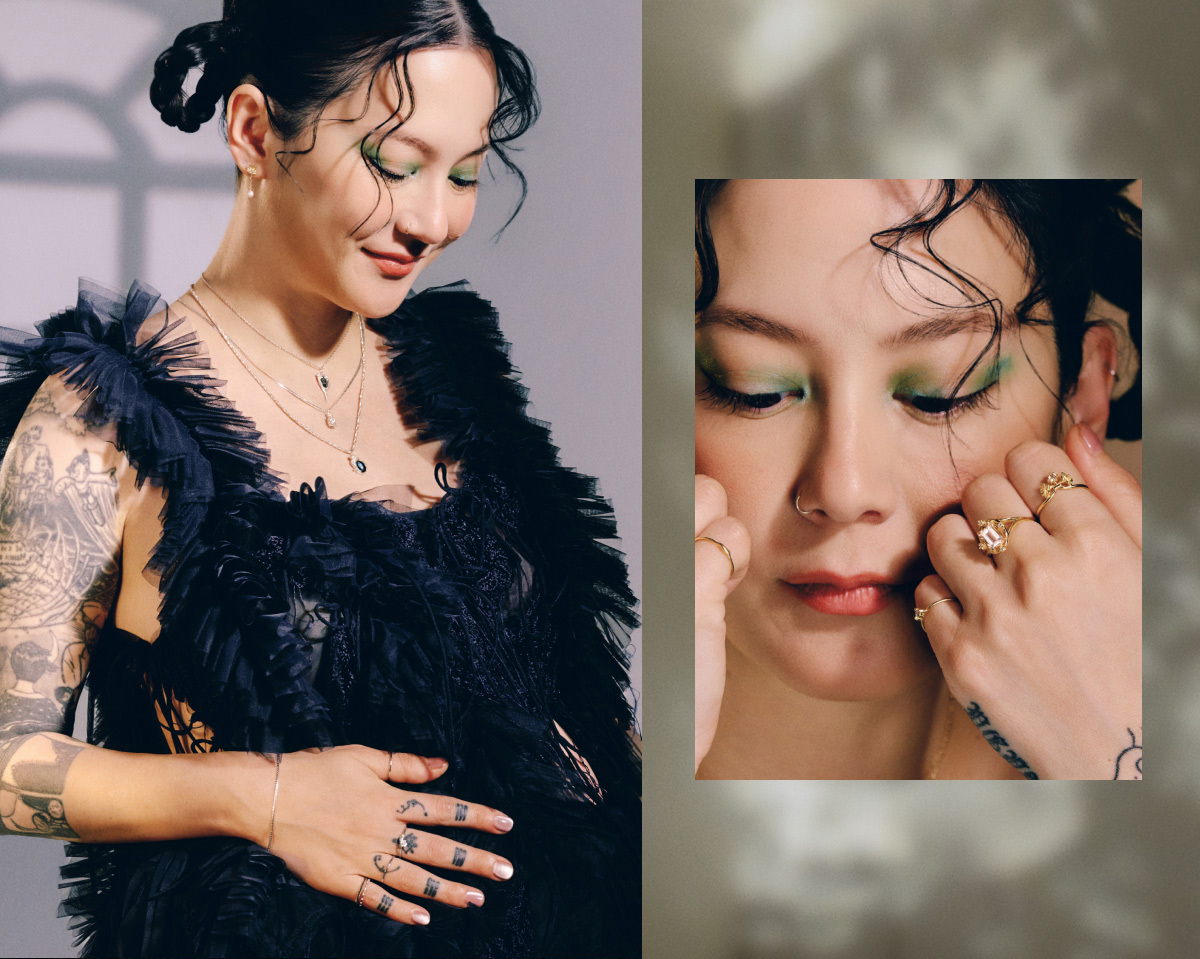 Michelle Zauner in a black ruffled outfit with green eyeshadow smiles gently, showing tattoos and layered necklaces. A close-up of her hands adorned with rings is also visible.