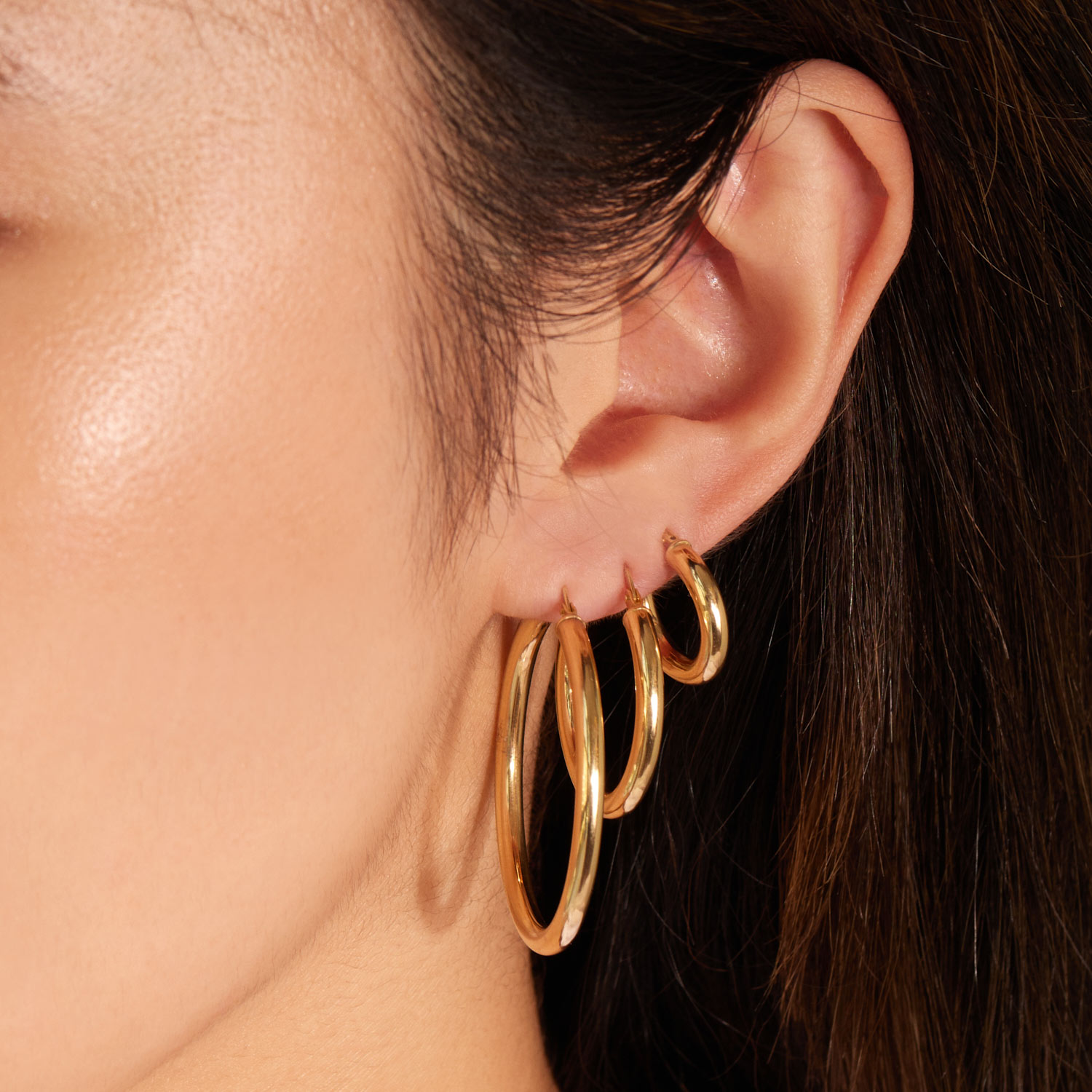 Three gold hoop earrings of varying sizes are arranged on a soft pink background. 