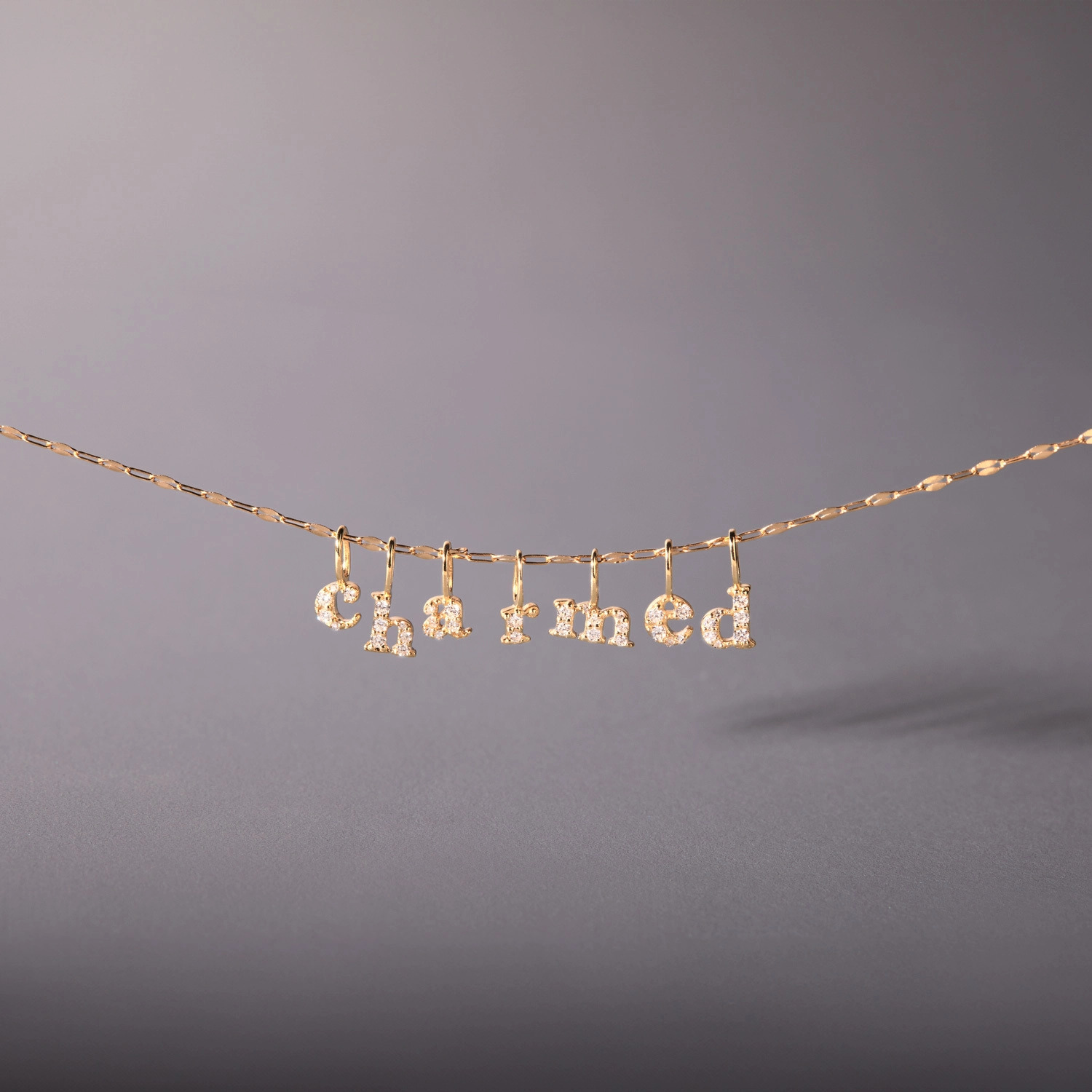 A delicate gold necklace with diamond-encrusted letters spelling "charmed" hangs elegantly.