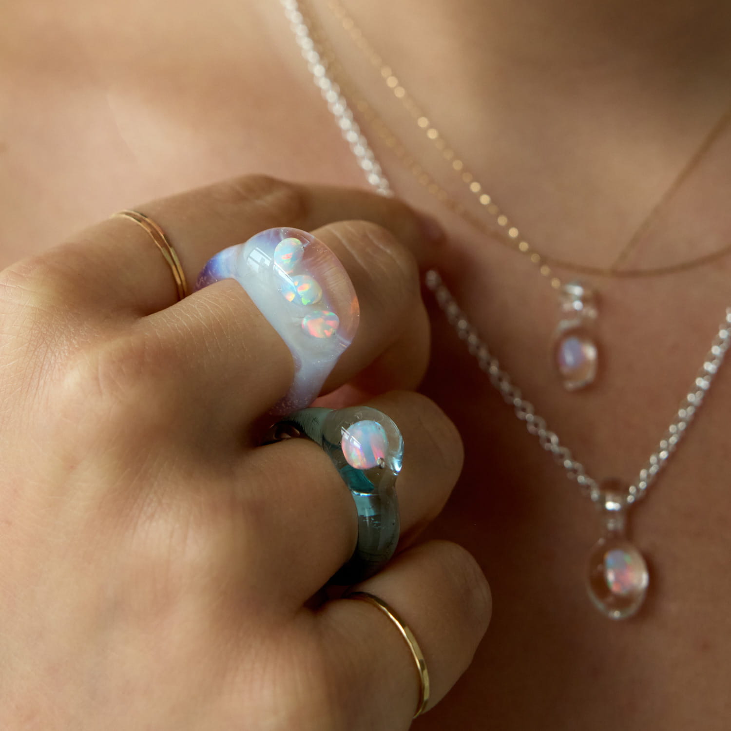 A close-up of a hand with three colorful rings and a silver necklace featuring an iridescent pendant.